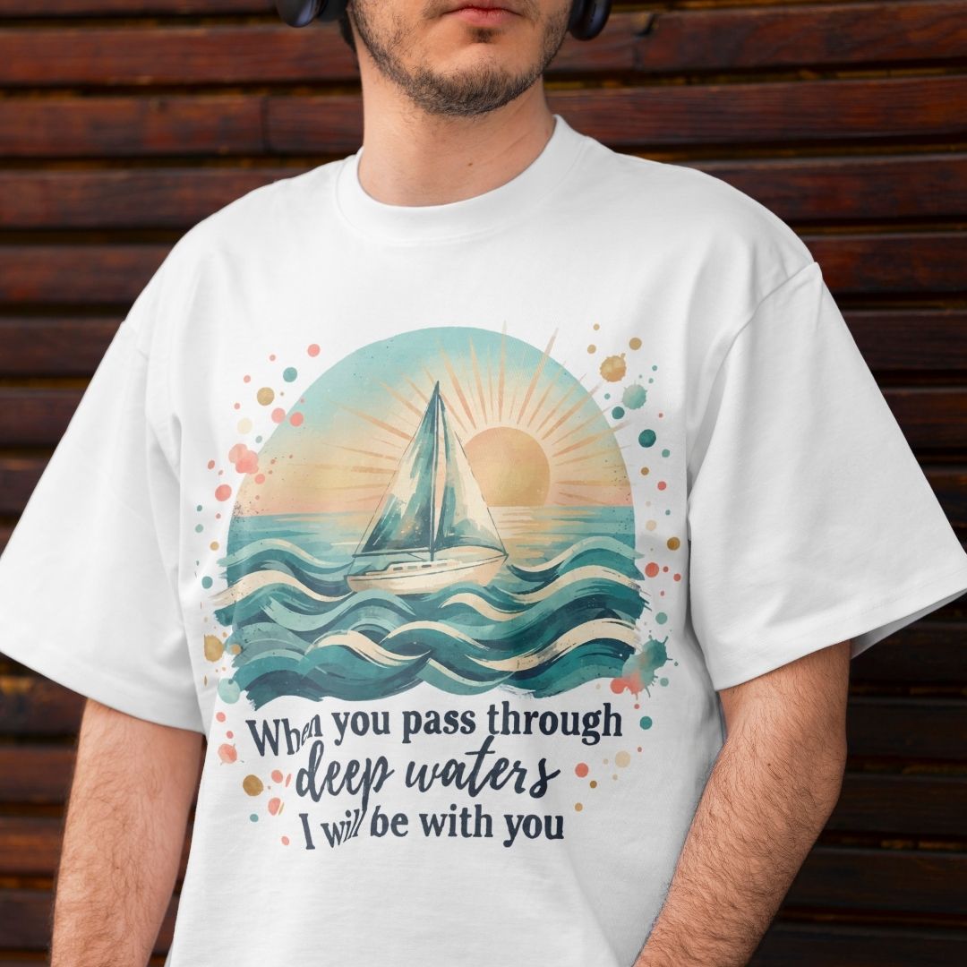 A man wearing a white graphic t-shirt with a watercolor sailboat design and the quote 'What you pass through deep waters I will be with you' against a dark wooden background.