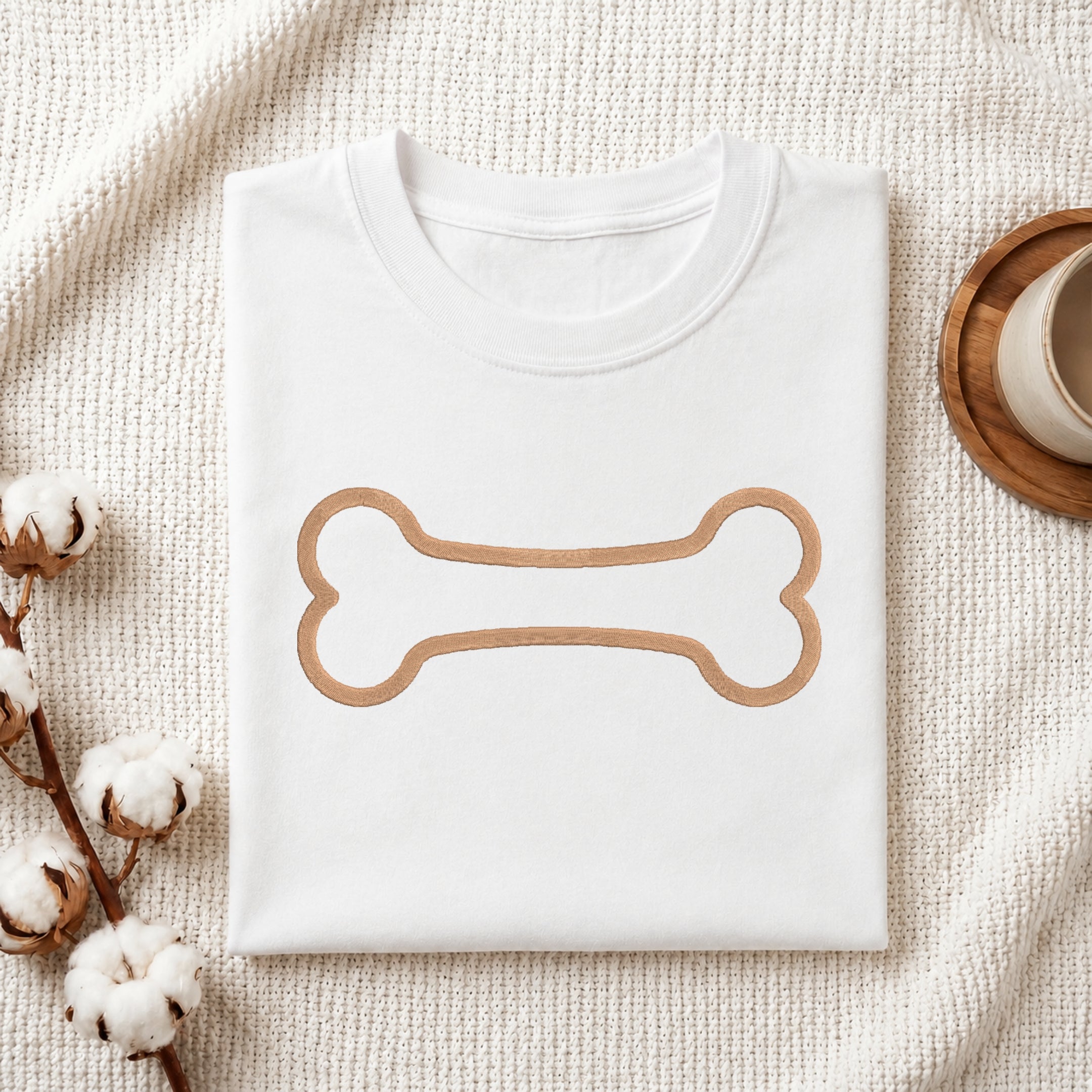 A folded white crewneck t-shirt with a tan bone outline graphic on the chest, styled on a textured cream knit blanket with cotton bolls and a wooden cup in the background.