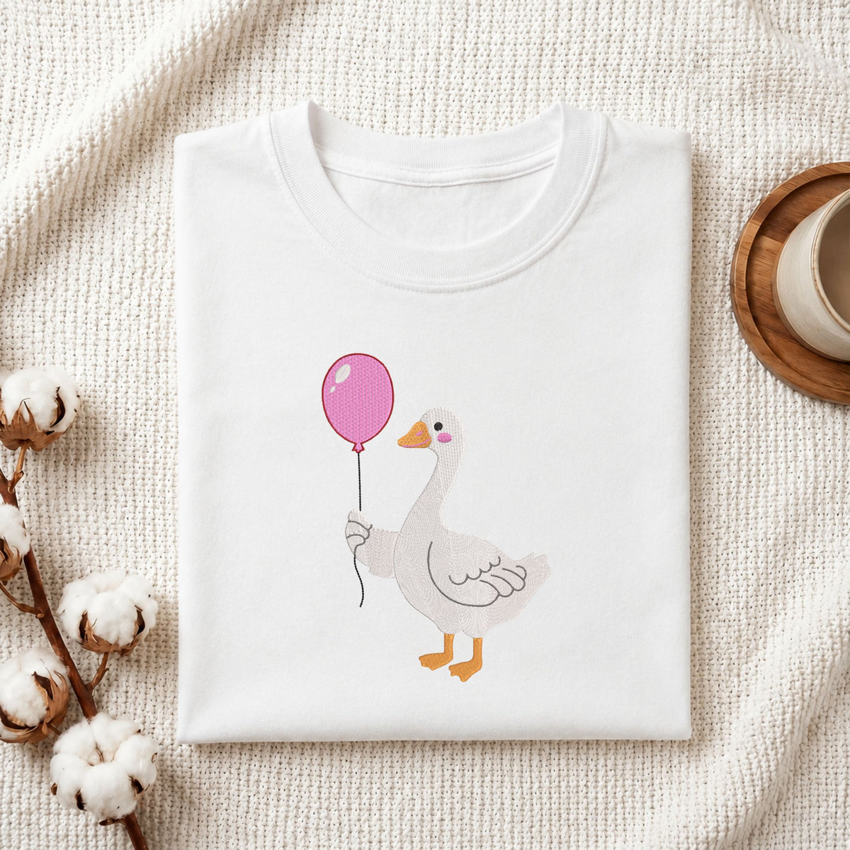  Folded white cotton t-shirt mockup featuring a cute duck wearing pink rain boots machine embroidery design, styled with cotton stems and a wooden cup   