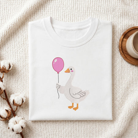  Folded white cotton t-shirt mockup featuring a cute duck wearing pink rain boots machine embroidery design, styled with cotton stems and a wooden cup   