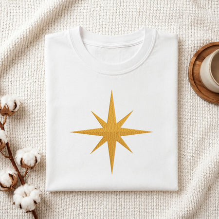 Folded white cotton t-shirt mockup featuring a golden starburst machine embroidery design, styled with cotton stems and a wooden cup