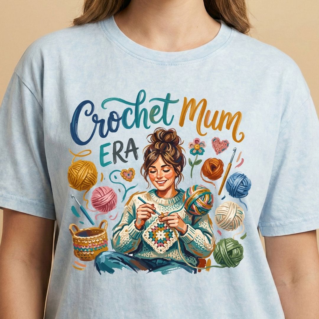 Light blue t-shirt with a colorful graphic of a woman crocheting and text 'Crochet Mum Era' on a beige background.