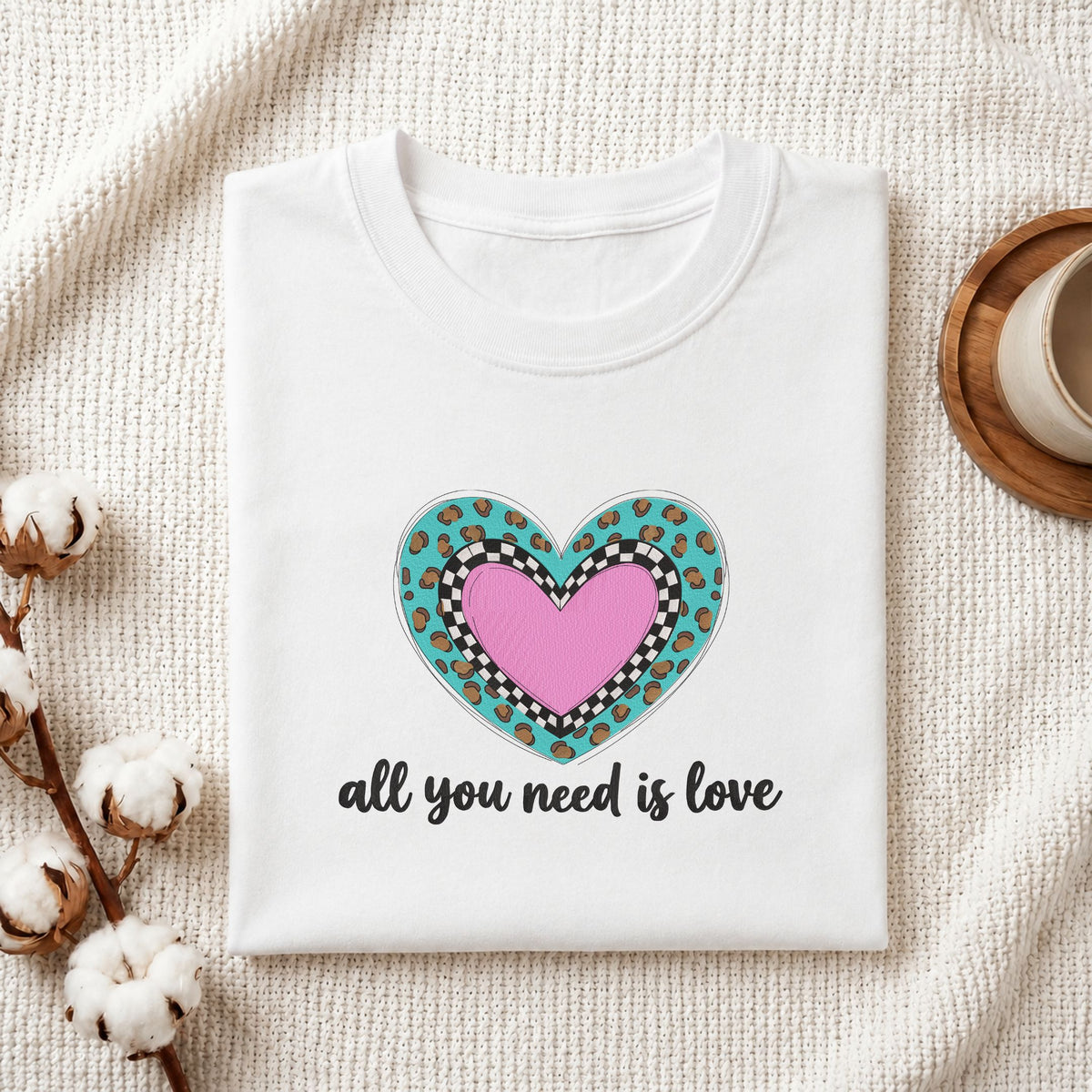 Folded white cotton t-shirt mockup featuring an "all you need is love" leopard heart machine embroidery design, styled with cotton stems and a wooden cup