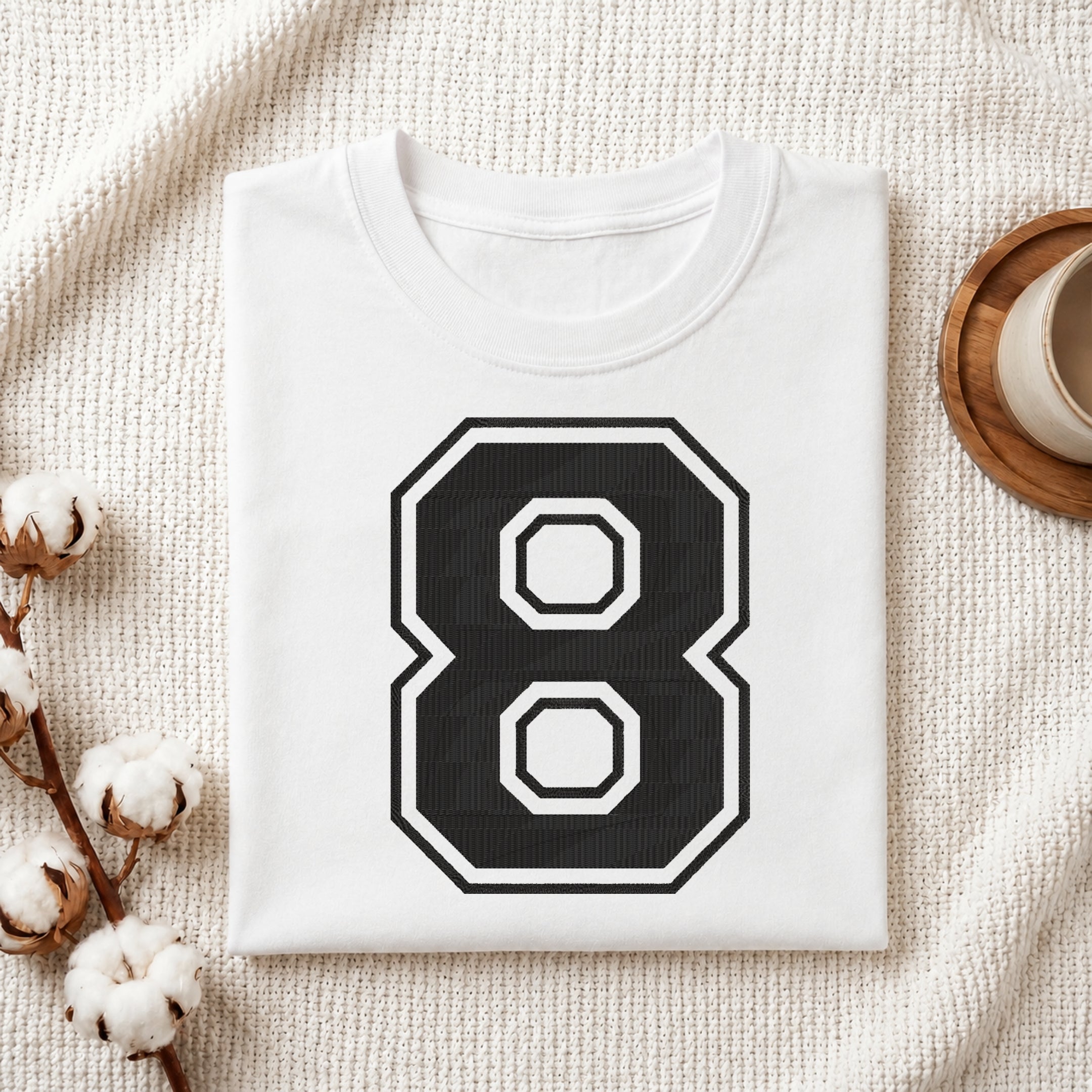 A folded white t-shirt with a large black and white number 8 graphic on the front, styled on a textured cream fabric with cotton stems and a wooden cup in the background.