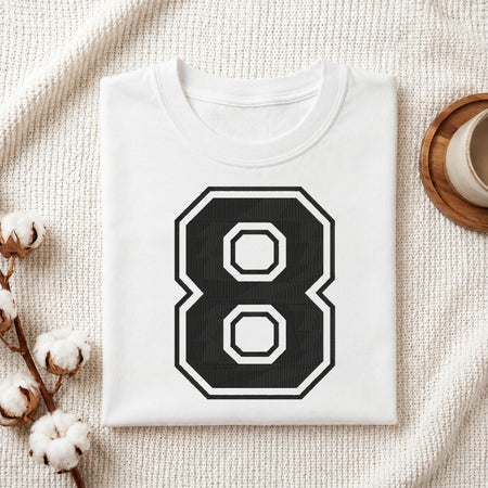 A folded white t-shirt with a large black and white number 8 graphic on the front, styled on a textured cream fabric with cotton stems and a wooden cup in the background.