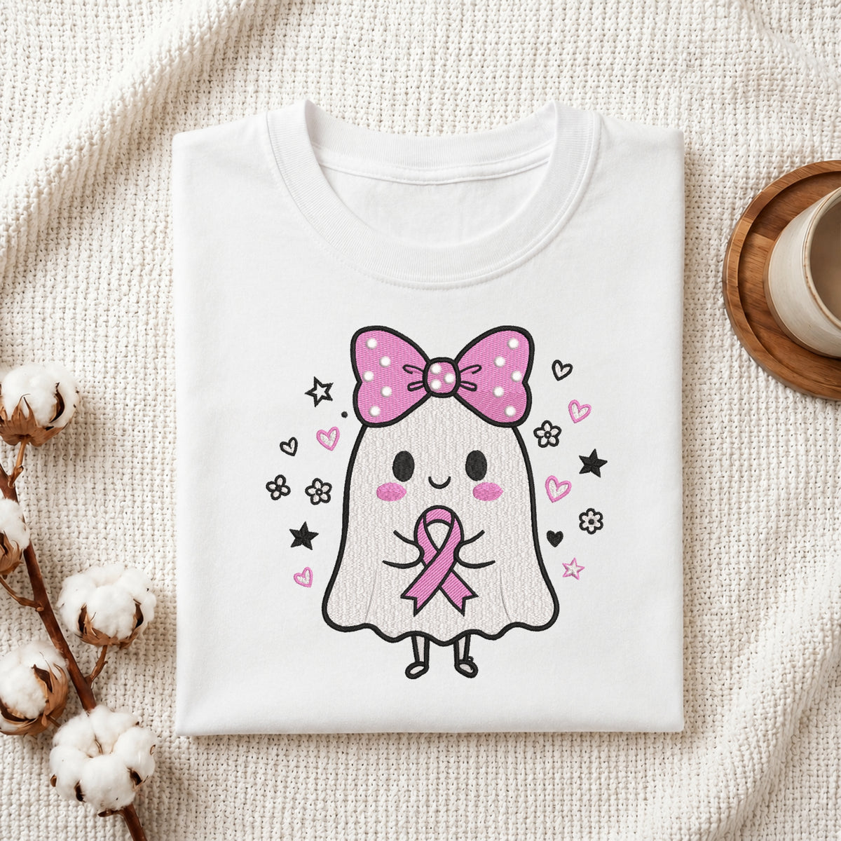 A neatly folded white t-shirt featuring the embroidered ghost holding a pink ribbon graphic, displayed on a textured white knit background with cotton stems and a wooden cup.