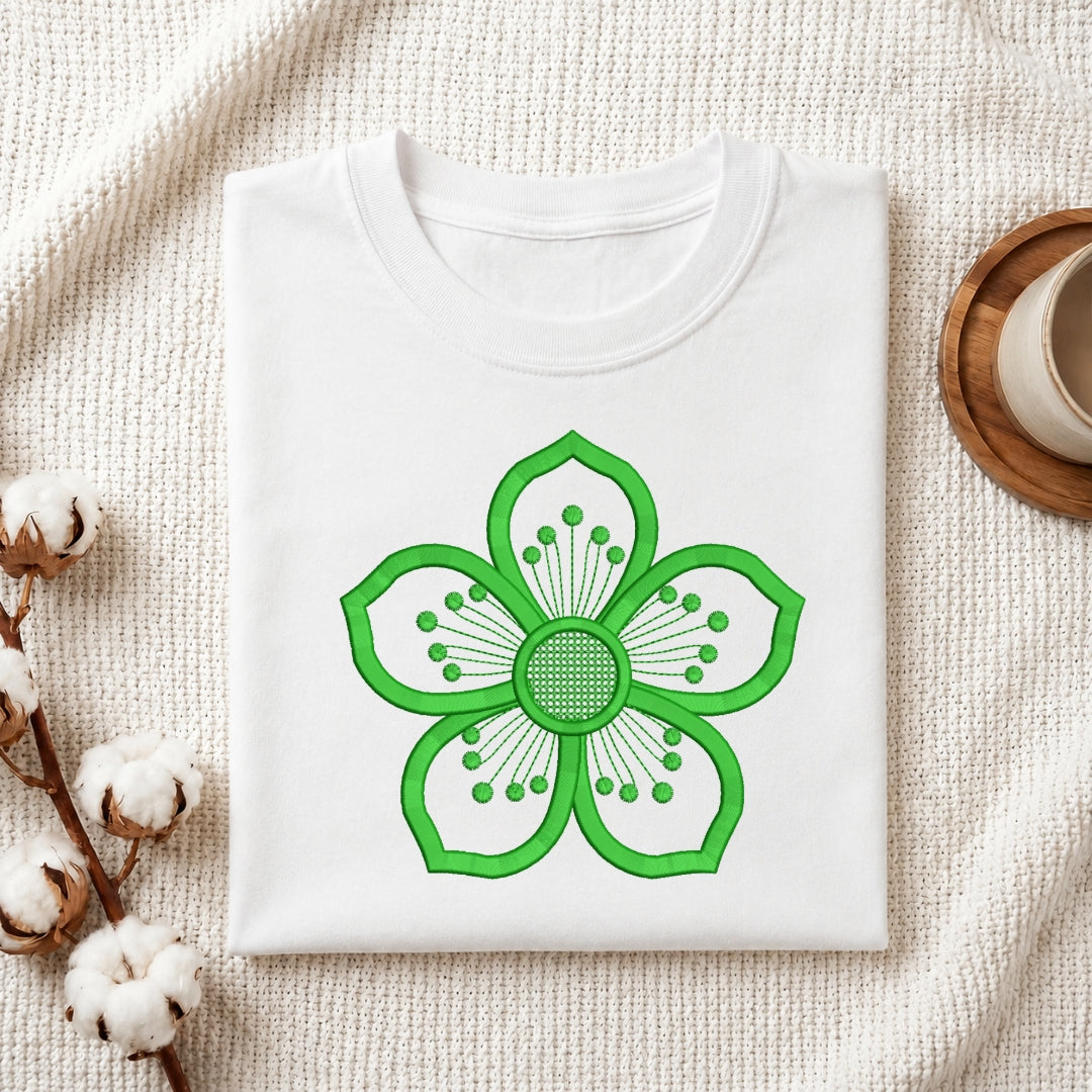 A folded white crewneck t-shirt with a bright green stylized flower graphic on the front, displayed on a textured cream knit blanket with cotton bolls and a wooden cup in the background.