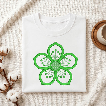 A folded white crewneck t-shirt with a bright green stylized flower graphic on the front, displayed on a textured cream knit blanket with cotton bolls and a wooden cup in the background.