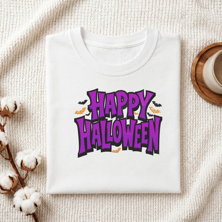 A folded white t-shirt with a 'Happy Halloween' graphic print in purple and orange, styled on a textured cream fabric with cotton stems and a small cup.