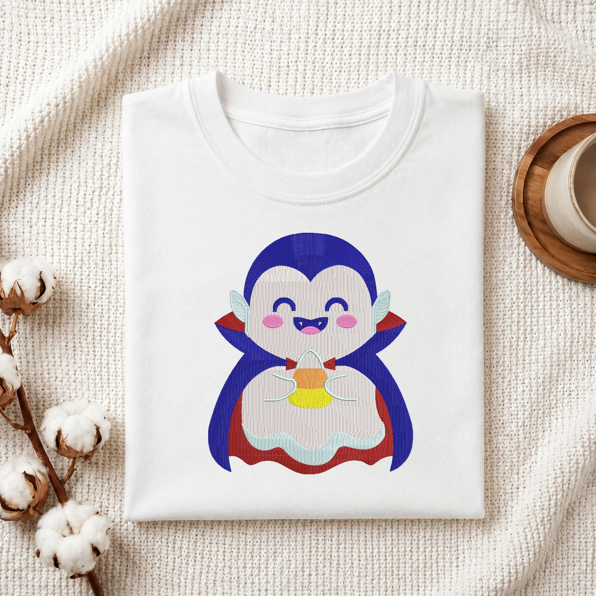 A folded white crewneck t-shirt featuring a cute cartoon penguin graphic, styled in a flat-lay on a textured cream fabric with cotton stems and a wooden cup on a coaster.