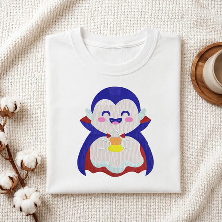 A folded white crewneck t-shirt featuring a cute cartoon penguin graphic, styled in a flat-lay on a textured cream fabric with cotton stems and a wooden cup on a coaster.