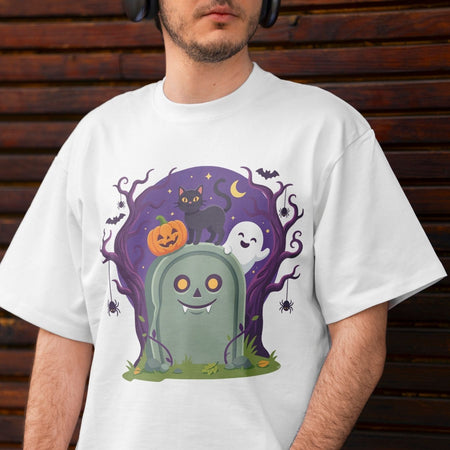 A man wearing a white crewneck t-shirt with a colorful Halloween graphic print featuring a tombstone, pumpkin, cat, ghost, and tree against a dark purple sky, set against a dark wooden background.
