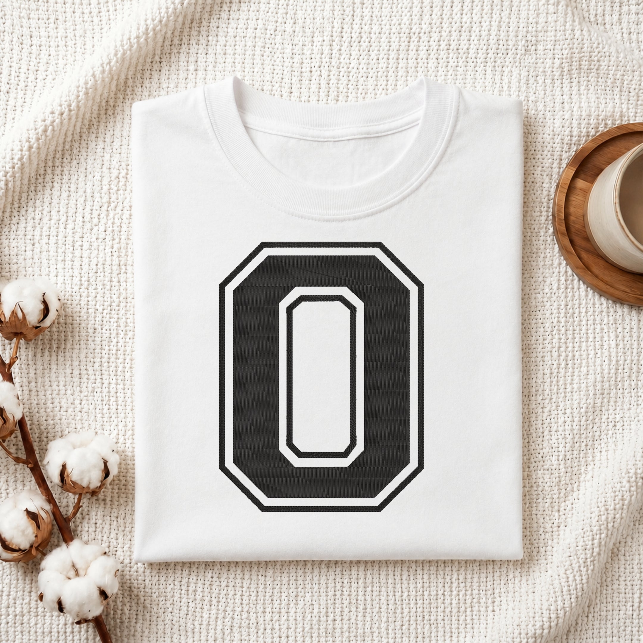 A folded white t-shirt with a large black and white number 0 graphic on the front, styled on a textured cream blanket with cotton bolls and a cup in the background.