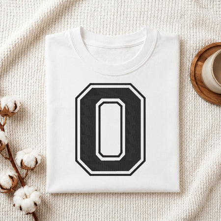 A folded white t-shirt with a large black and white number 0 graphic on the front, styled on a textured cream blanket with cotton bolls and a cup in the background.