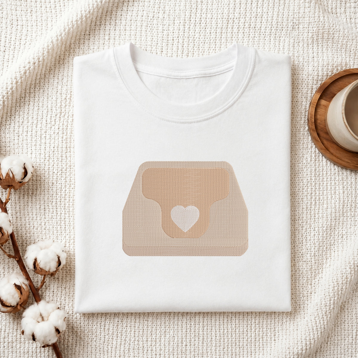 A neatly folded white crewneck t-shirt featuring a stitched heart purse design, styled with cotton branches and a wooden coaster.