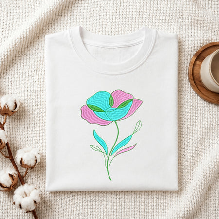A folded white crewneck t-shirt with a colorful embroidered flower design, displayed on a textured cream fabric background with cotton bolls and a wooden cup in the corners.