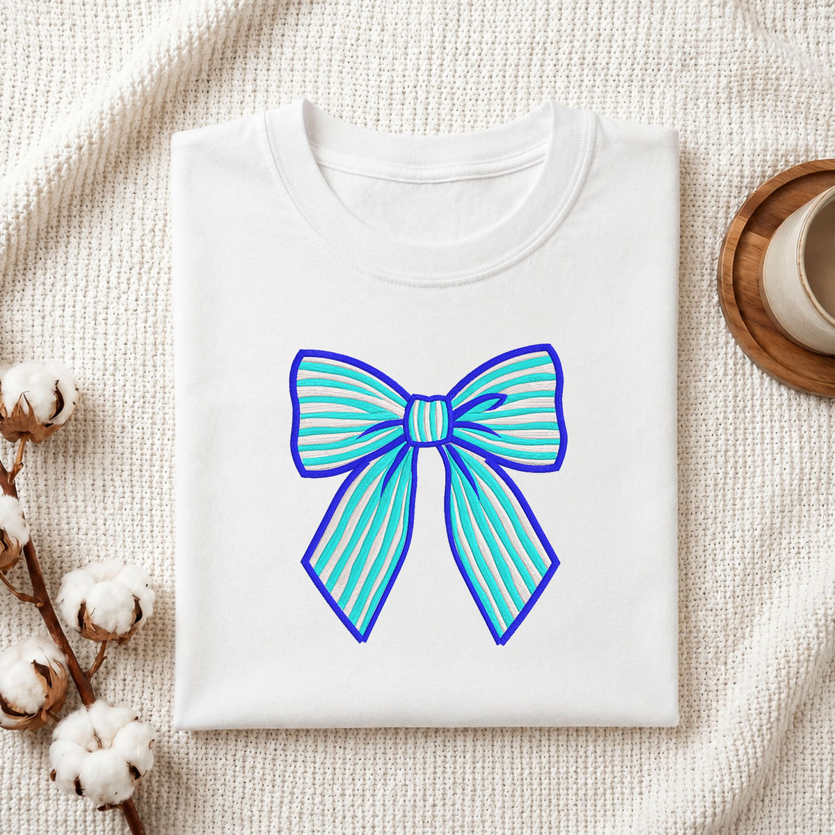 A folded white crewneck t-shirt with a blue and light blue striped bow graphic on the front, styled on a textured cream knit blanket with cotton bolls and a wooden cup in the background.