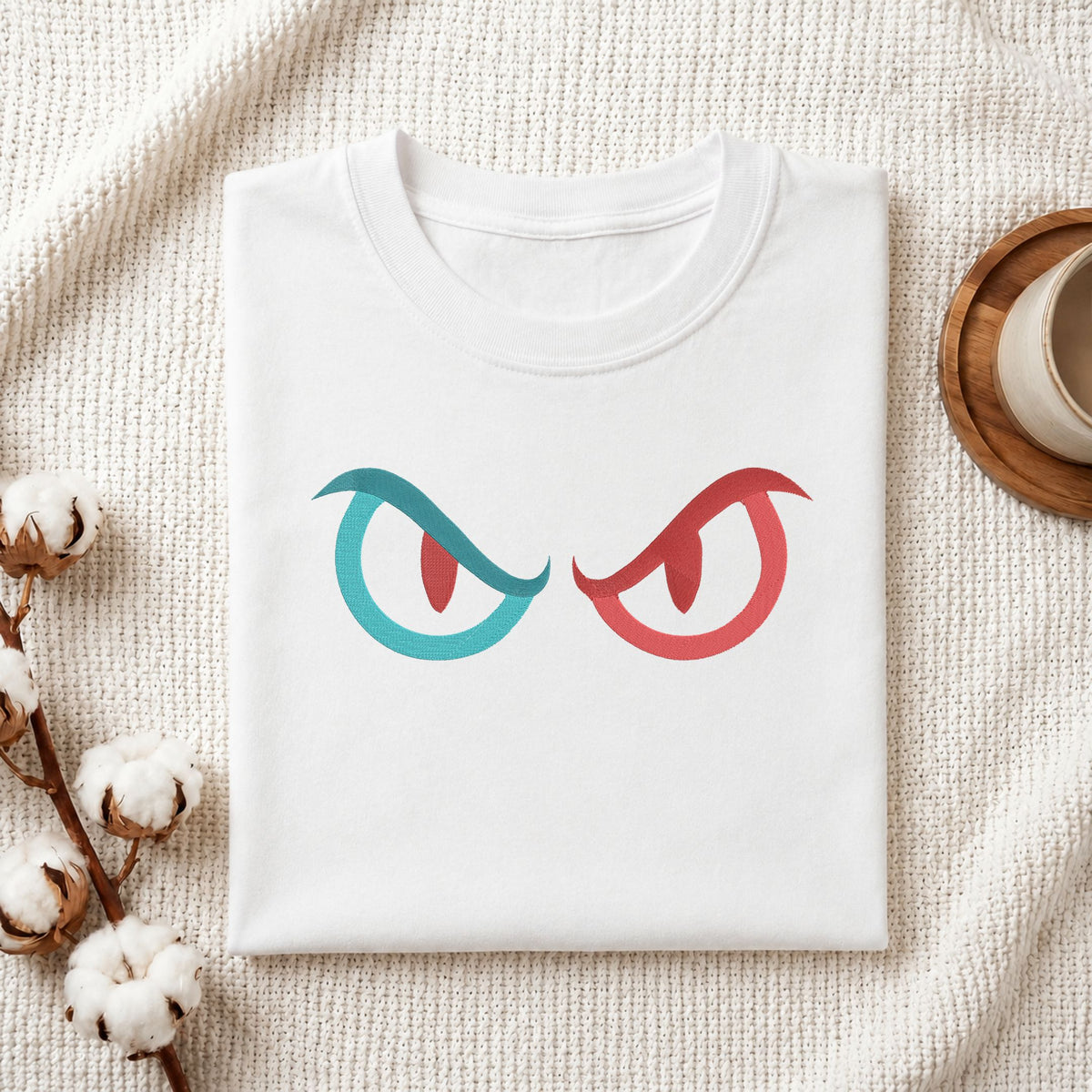 A folded white t-shirt with a graphic of two angry eyes, one in teal and one in red, displayed on a textured cream knit blanket with cotton bolls and a cup on a wooden tray.