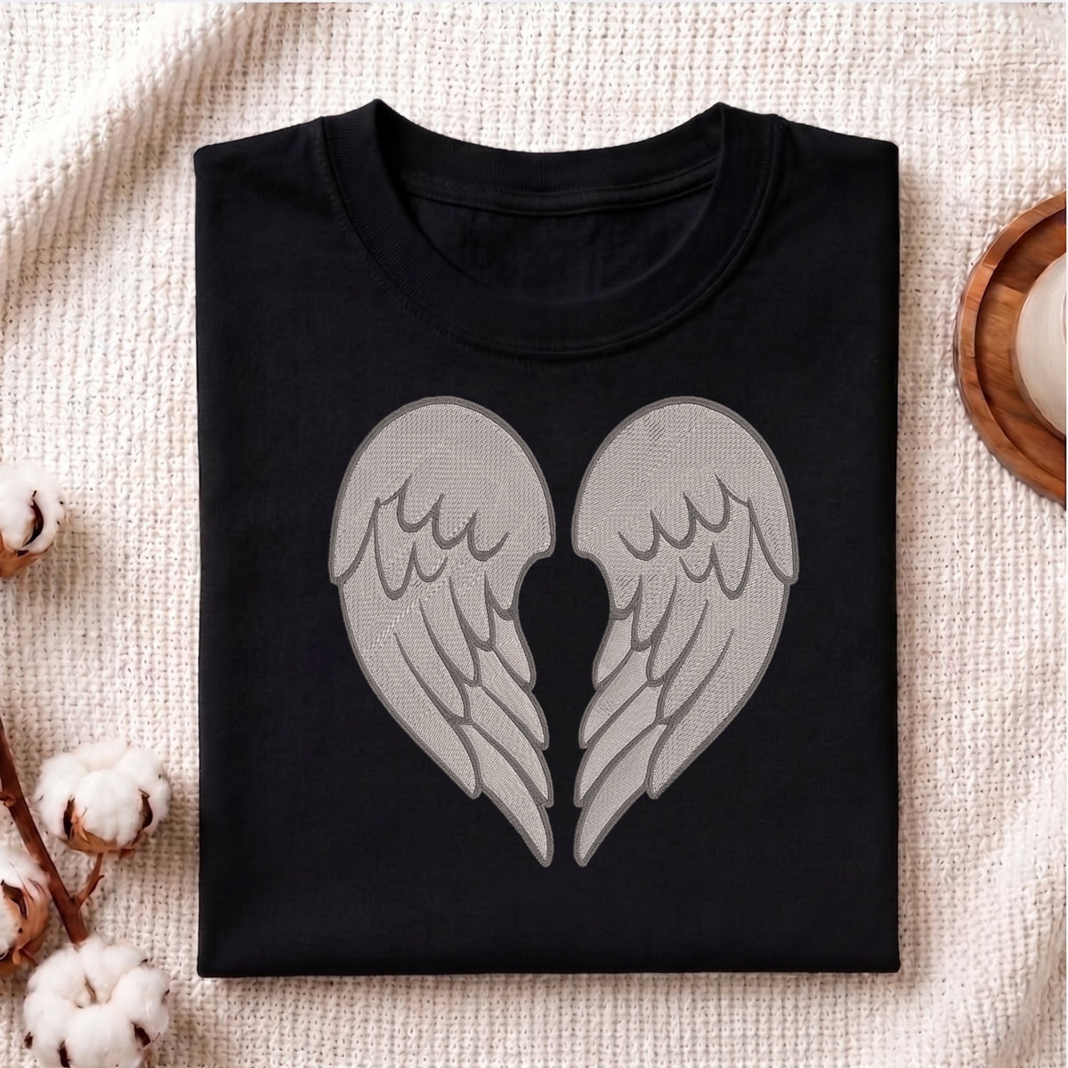 A folded black crewneck t-shirt featuring a large graphic of two gray angel wings that form a heart shape in the center, displayed on a textured beige fabric background with cotton bolls and a wooden coaster.