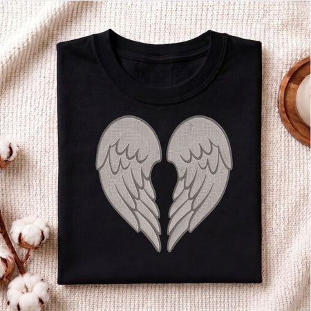 A folded black crewneck t-shirt featuring a large graphic of two gray angel wings that form a heart shape in the center, displayed on a textured beige fabric background with cotton bolls and a wooden coaster.