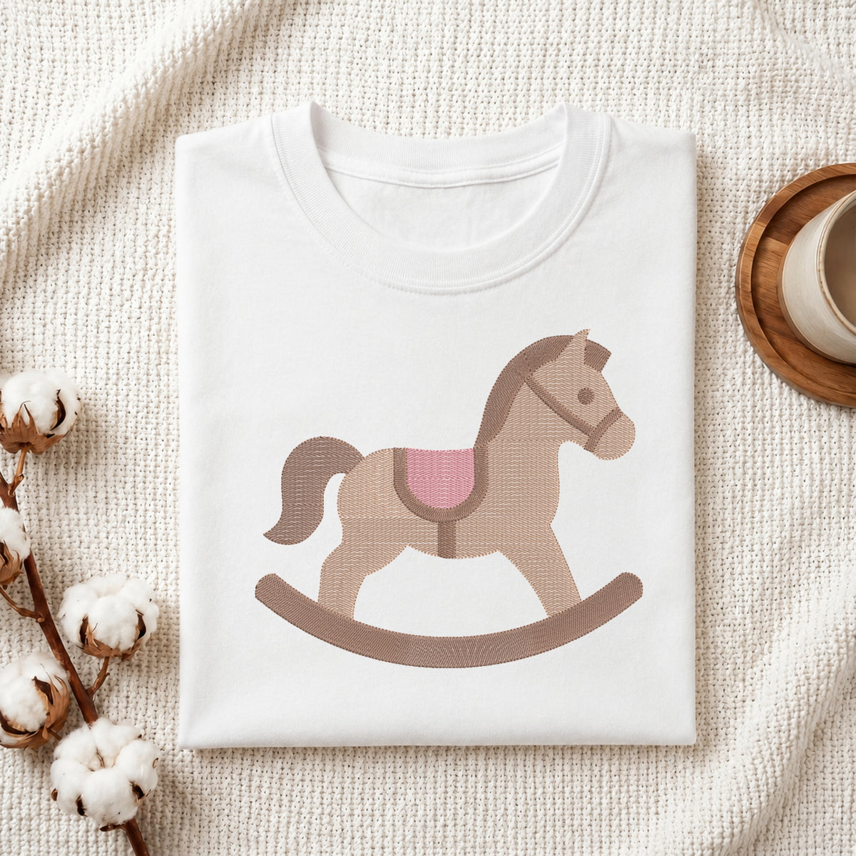 A folded white T-shirt with a stylized rocking horse graphic in tan, brown, and pink, displayed on a textured cream fabric with cotton stems and a small cup in the corners.