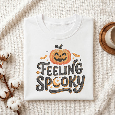 A folded white t-shirt with a 'FEELING SPOOKY' graphic featuring a jack-o'-lantern and bats, styled on a textured cream blanket with cotton bolls and a mug.