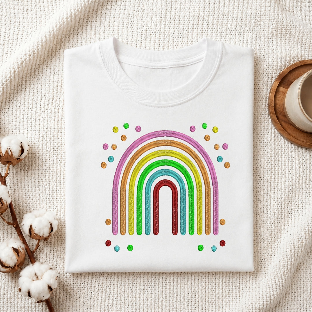 A folded white crewneck t-shirt featuring a colorful rainbow graphic with small multicolored dots, styled on a textured cream fabric background with cotton bolls and a wooden cup.