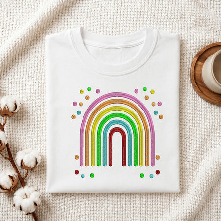 A folded white crewneck t-shirt featuring a colorful rainbow graphic with small multicolored dots, styled on a textured cream fabric background with cotton bolls and a wooden cup.