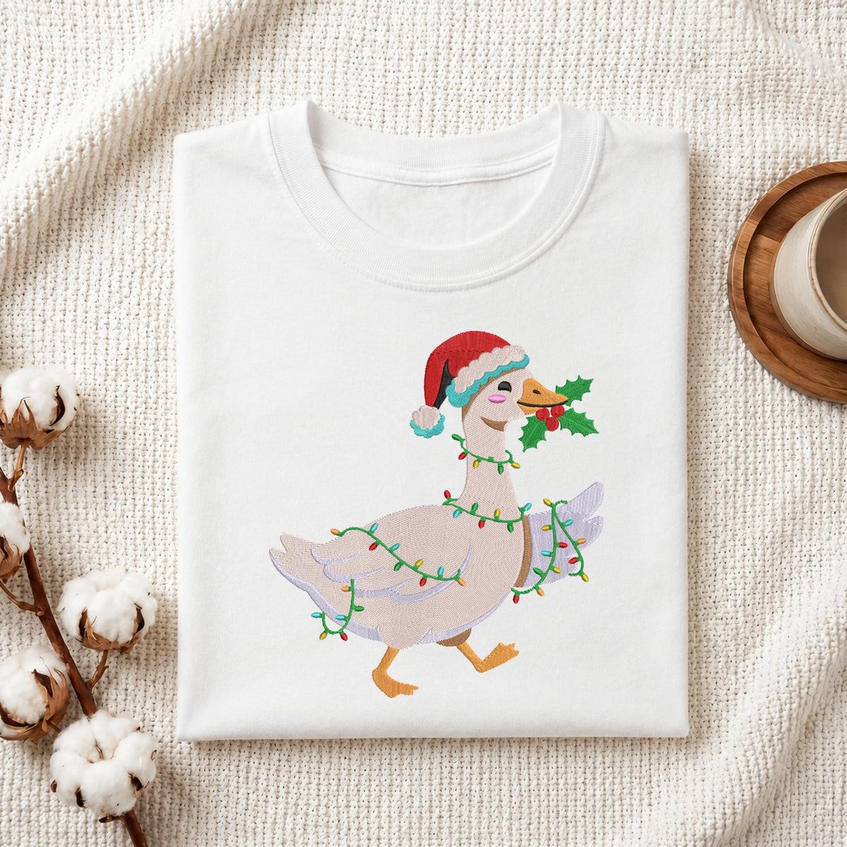 A folded white crewneck t-shirt with a graphic of a goose wearing a red Santa hat and holiday lights, styled on a textured cream fabric with cotton bolls and a mug.