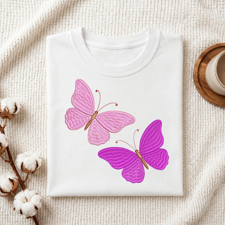 A folded white crewneck t-shirt featuring two embroidered butterflies, one pink and one purple, displayed on a textured cream fabric background with decorative cotton bolls and a wooden cup.