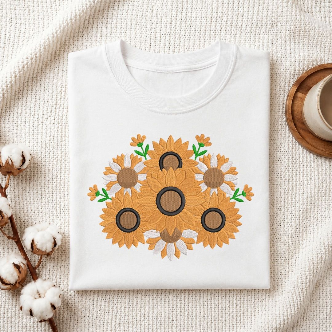 A folded white crewneck t-shirt featuring a graphic print of sunflowers, styled on a textured cream knit blanket with cotton bolls and a wooden cup in the background.