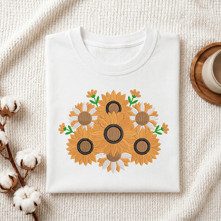 A folded white crewneck t-shirt featuring a graphic print of sunflowers, styled on a textured cream knit blanket with cotton bolls and a wooden cup in the background.