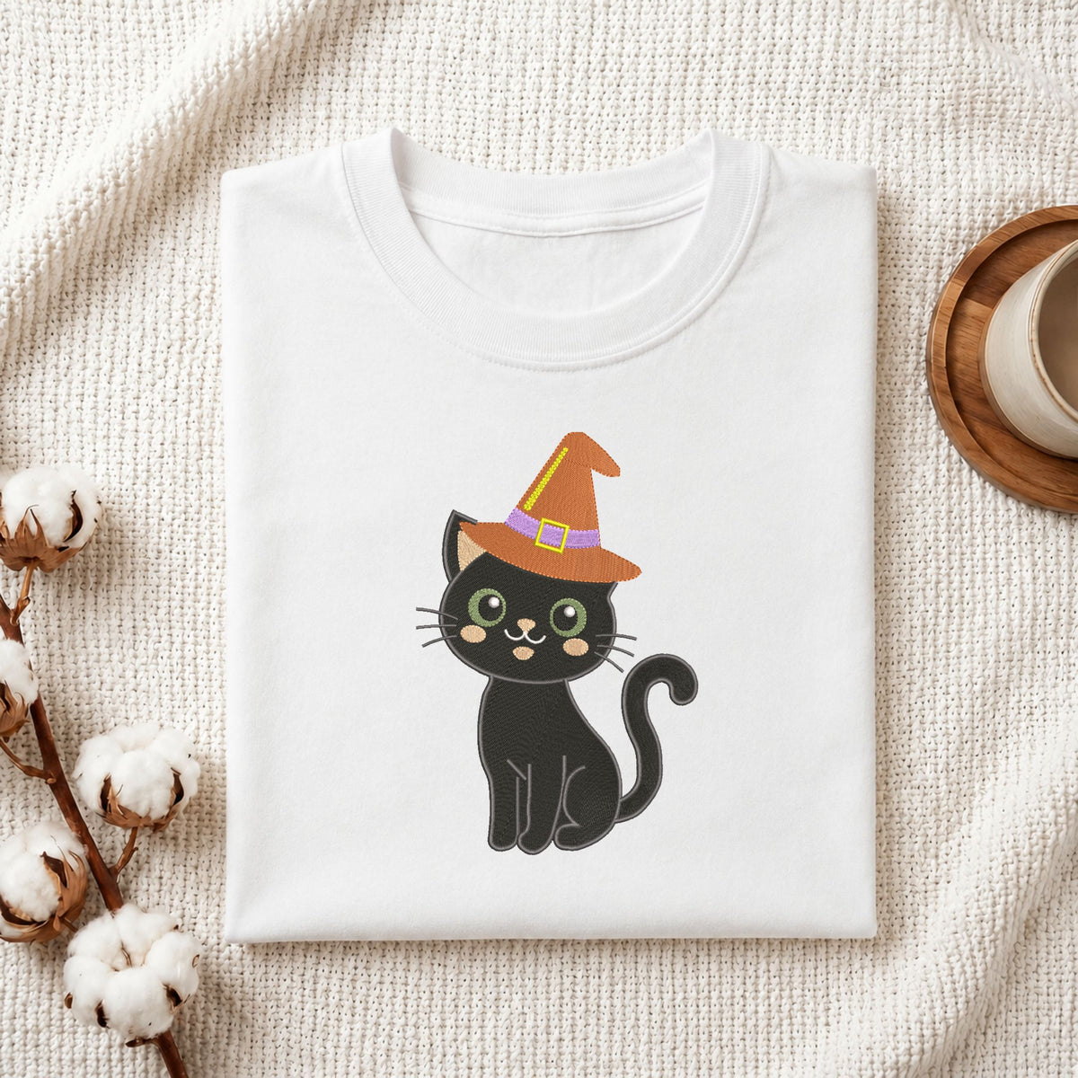 Folded white cotton t-shirt mockup featuring a black cat in a witch hat machine embroidery design, styled with cotton stems and a wooden cup.