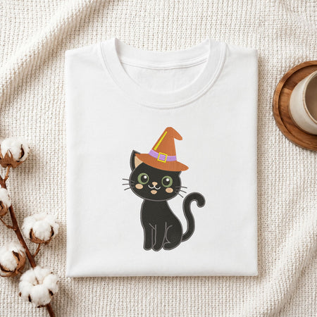 Folded white cotton t-shirt mockup featuring a black cat in a witch hat machine embroidery design, styled with cotton stems and a wooden cup.