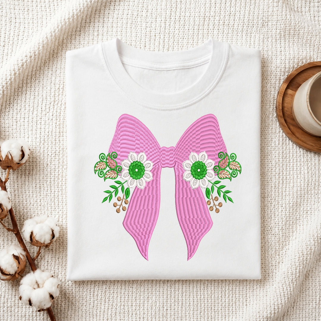 A folded white crewneck t-shirt displayed on a textured cream fabric background, decorated with cotton bolls on the left and a wooden cup on the right. The t-shirt features a large pink striped bow graphic with green and white floral details.