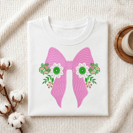 A folded white crewneck t-shirt displayed on a textured cream fabric background, decorated with cotton bolls on the left and a wooden cup on the right. The t-shirt features a large pink striped bow graphic with green and white floral details.