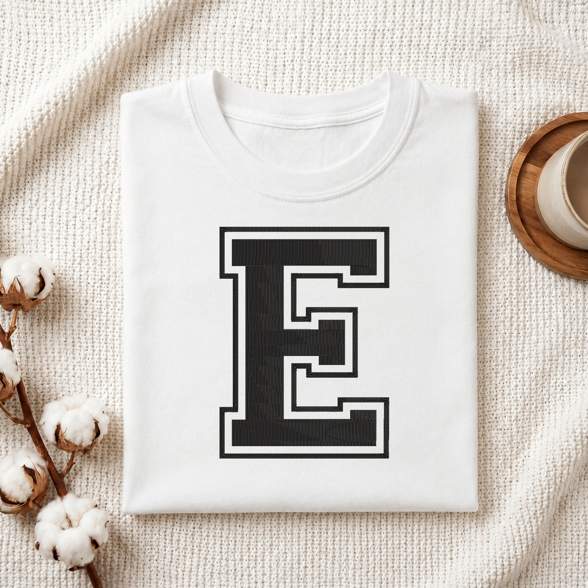 A folded white t-shirt with a large black block letter 'E' outlined in white on the front, styled on a textured cream fabric with cotton stems and a small cup in the background.