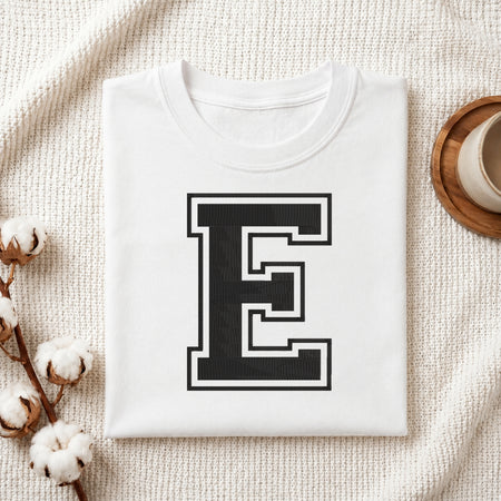 A folded white t-shirt with a large black block letter 'E' outlined in white on the front, styled on a textured cream fabric with cotton stems and a small cup in the background.