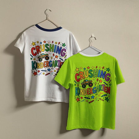 Two children's short-sleeve T-shirts hanging on hangers against a plain beige wall. The shirt on the left is white with a dark navy collar and shows a colorful back graphic reading 'CRUSHING KINDERGARTEN' with stars, crayons, and cartoon vehicles.