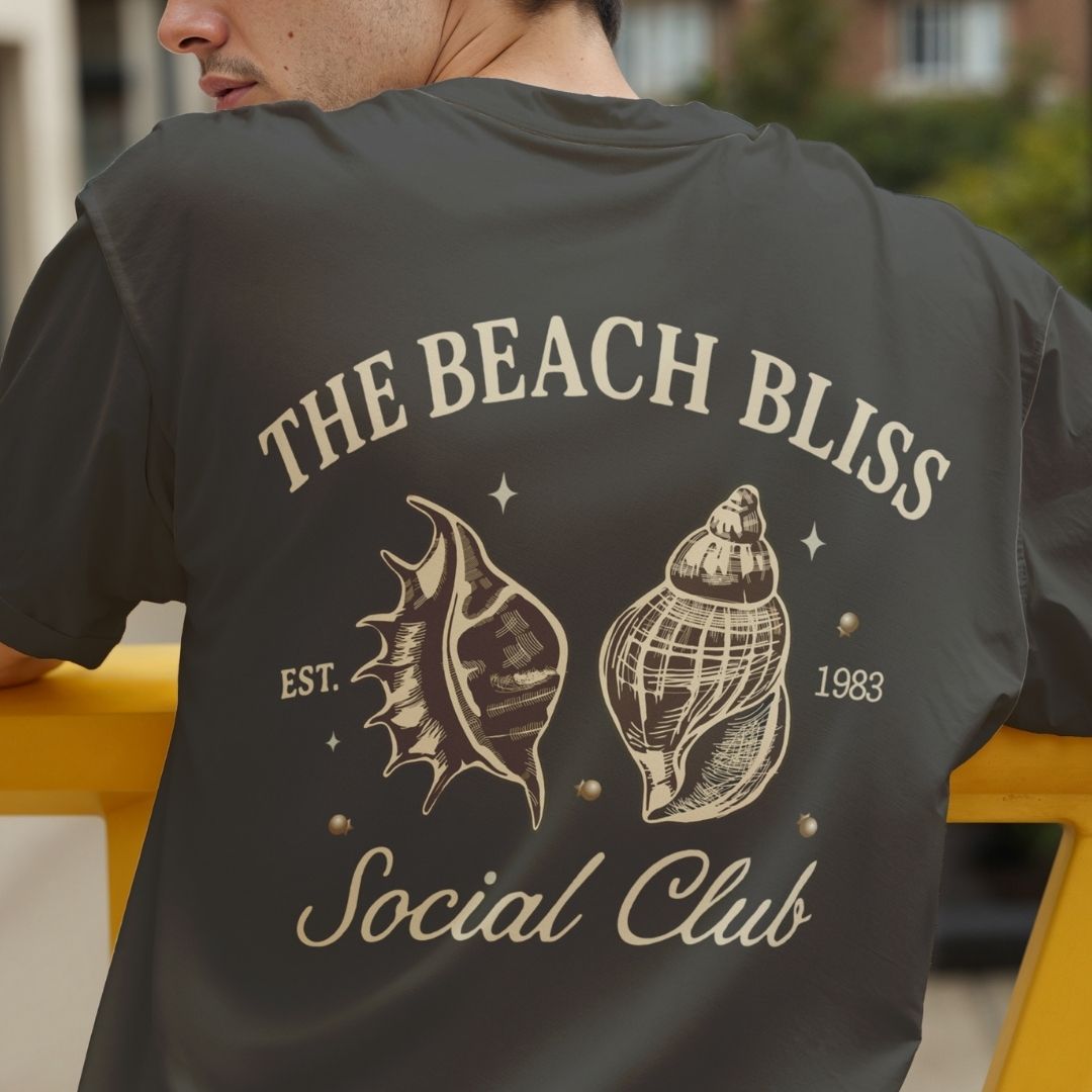 Close up of the back of a man's charcoal grey t-shirt showing a large retro coastal style design with cream-colored sketches of seashells and text reading The Beach Bliss Social Club.