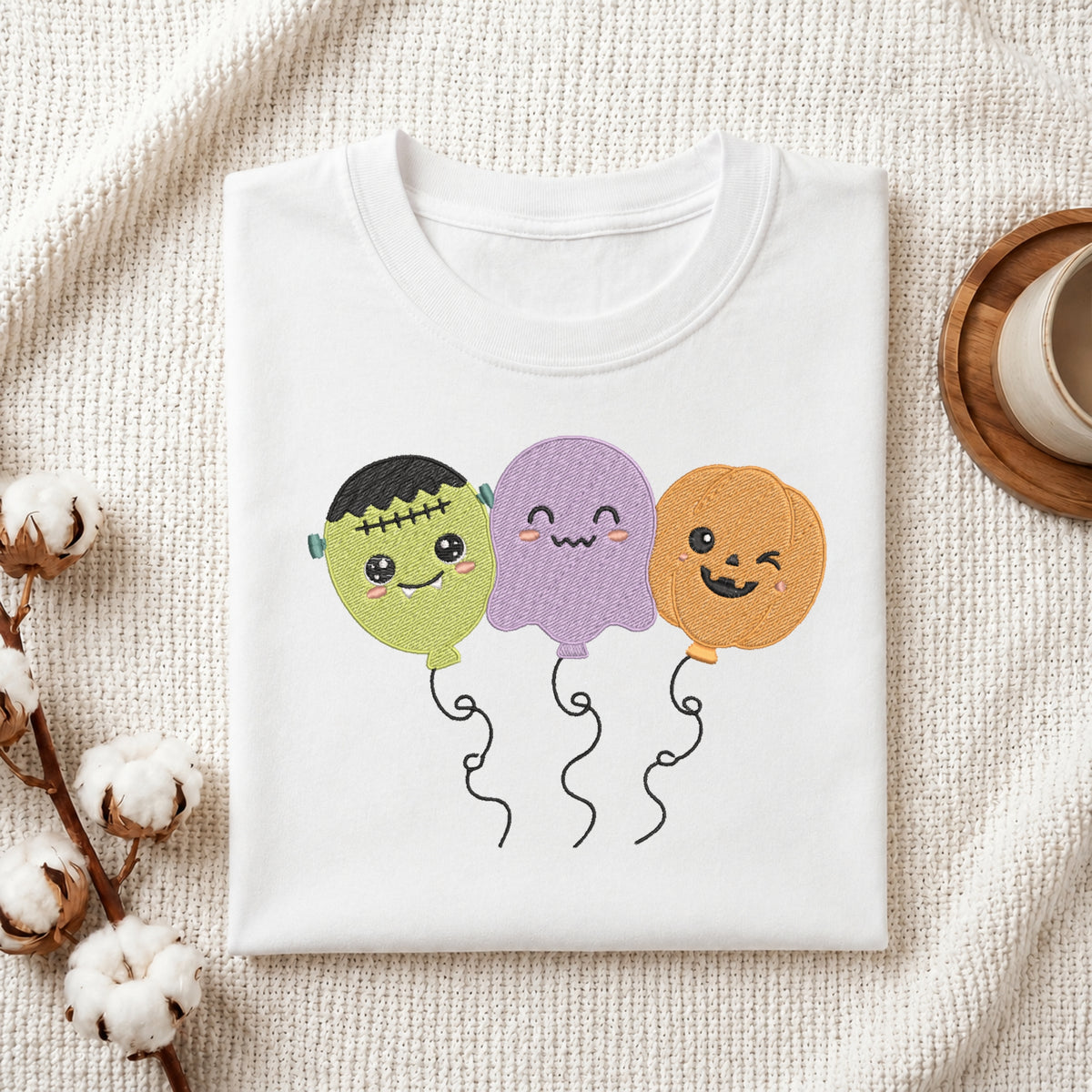 Folded white t-shirt mockup with a cute Halloween balloon trio embroidery design showing Frankenstein, a ghost, and a pumpkin.