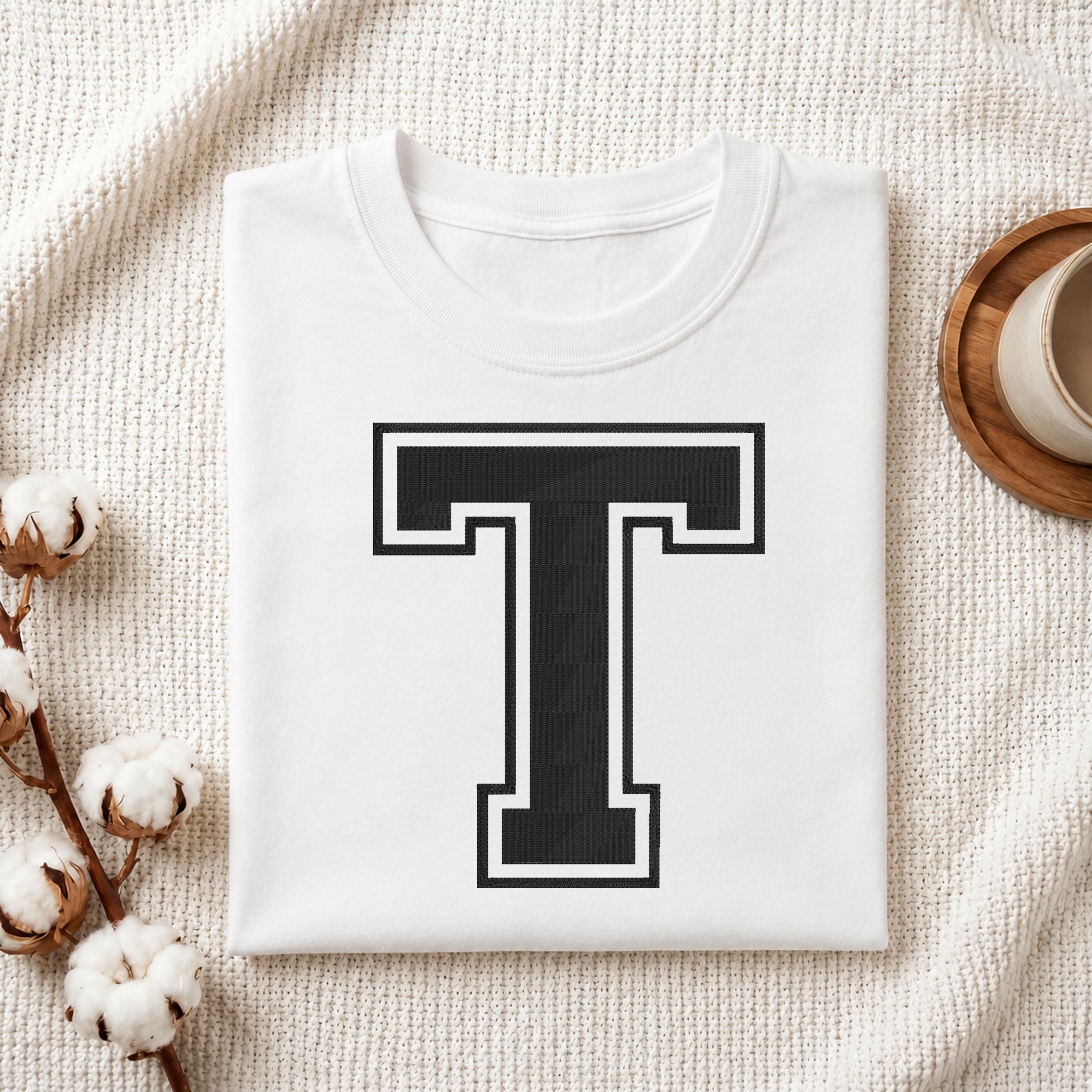 A folded white crewneck t-shirt with a large black and white block letter 'T' printed on the front, styled on a textured cream fabric with cotton bolls and a cup on a wooden coaster.