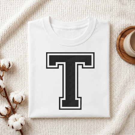 A folded white crewneck t-shirt with a large black and white block letter 'T' printed on the front, styled on a textured cream fabric with cotton bolls and a cup on a wooden coaster.