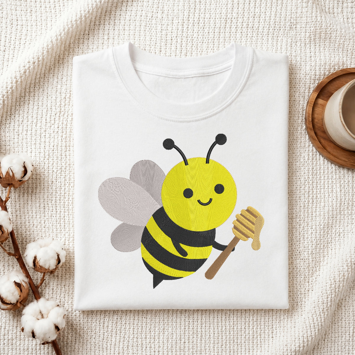 A folded white crewneck t-shirt featuring a cute cartoon bee graphic holding a honeycomb, styled on a textured cream fabric with cotton bolls and a wooden cup in the background.