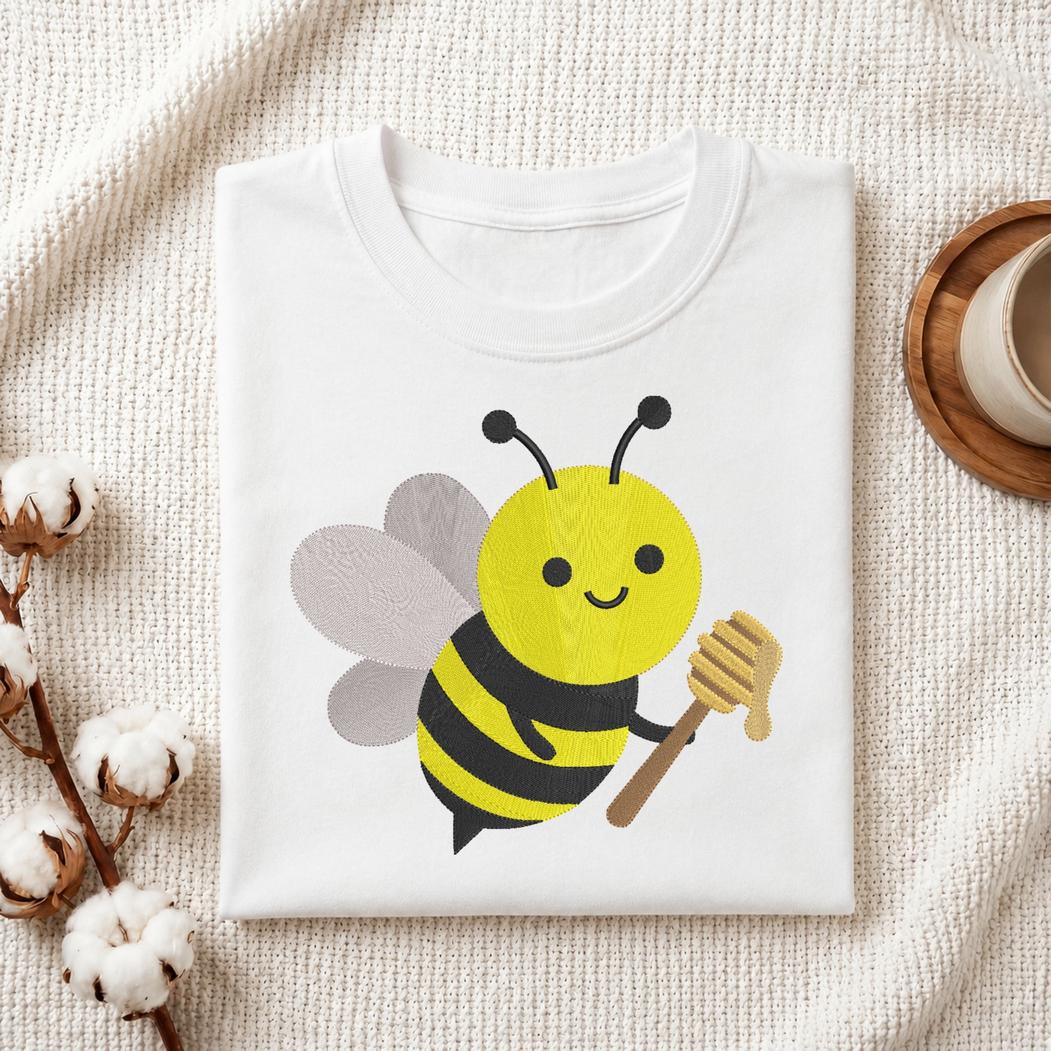 A folded white crewneck t-shirt featuring a cute cartoon bee graphic holding a honeycomb, styled on a textured cream fabric with cotton bolls and a wooden cup in the background.