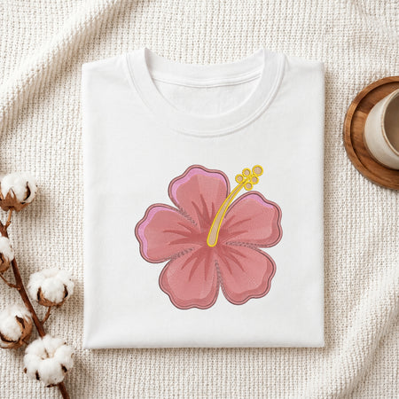 A folded white T-shirt with a large pink hibiscus flower graphic and yellow stamen detail on the front, styled on a textured cream fabric with cotton stems and a small cup in the corners.