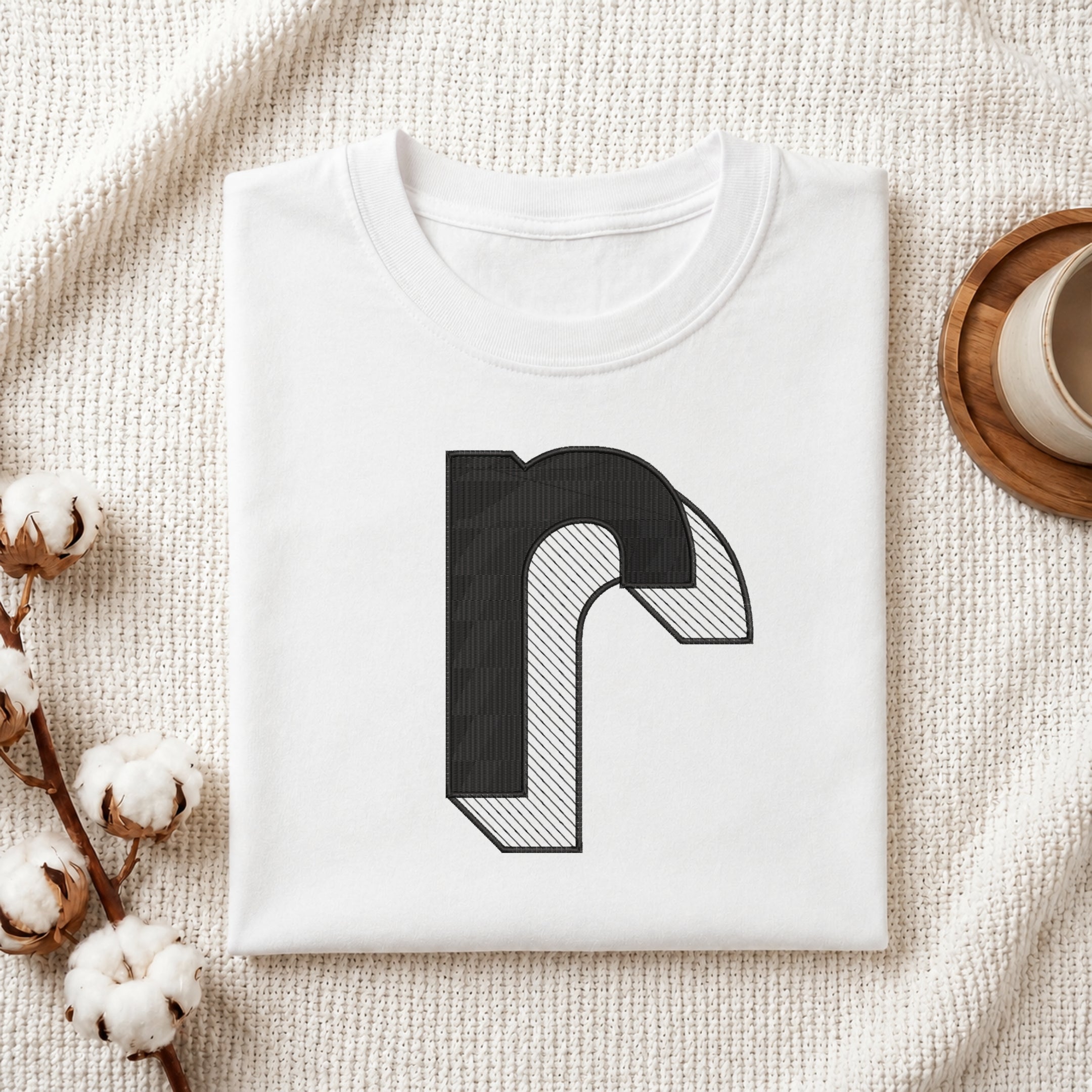 A folded white t-shirt with a black stylized letter 'r' graphic printed on the front, displayed on a textured cream knit blanket with decorative cotton bolls and a cup in the background.