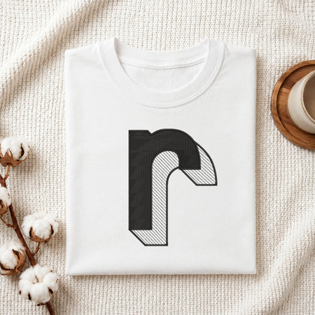 A folded white t-shirt with a black stylized letter 'r' graphic printed on the front, displayed on a textured cream knit blanket with decorative cotton bolls and a cup in the background.