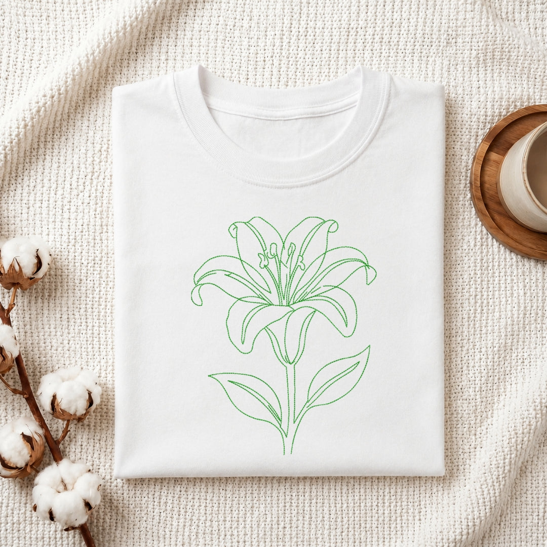 A folded white crewneck t-shirt with a green line art drawing of a lily flower on the front, styled on a textured cream fabric background with cotton bolls and a cup on a wooden coaster.