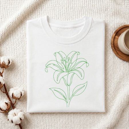 A folded white crewneck t-shirt with a green line art drawing of a lily flower on the front, styled on a textured cream fabric background with cotton bolls and a cup on a wooden coaster.